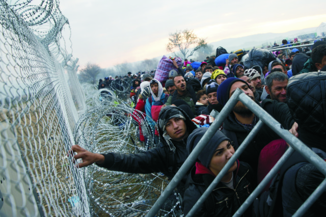 Na snímke migranti a utečenci čakajú pri plote na grécko-macedónskej hranici v snahe prejsť do Macedónska v Idomeni