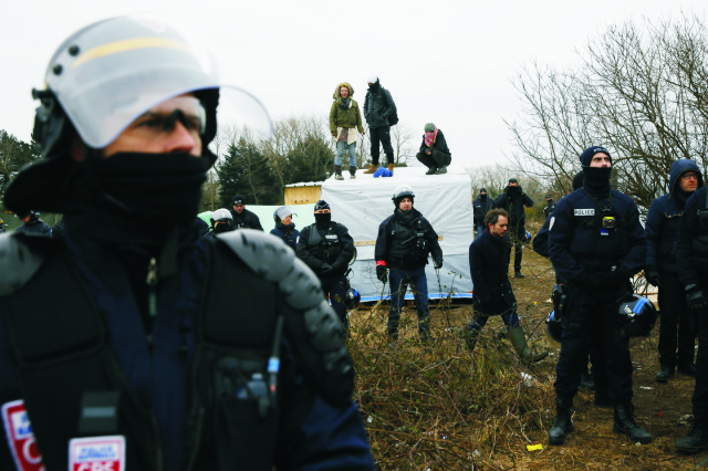 Policajti obklopujú migrantov a aktivistov stojacich na streche príbytkov v stanovom tábore pri francúzskom Calais 1. marca 2016 pred jeho zbúraním. Na predmestí Calais sa usadilo niekoľko tisícok afrických utečencov zo Stredomoria. Francúzske úrady začali odstraňovať rozsiahly tábor migrantov v meste Calais, kde žijú tisíce ľudí, dúfajúcich, že sa im podarí dostať do Británie za lepším životom